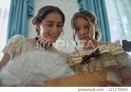 Low angle view of two romantic young ladies reading letter together and smiling in classic vintage setting 121270083