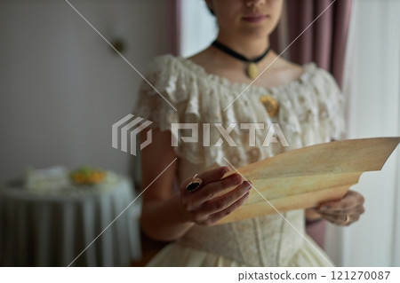 Close up of female hands holding letter in tinted parchment while standing by window in elegant vintage interior, copy space Close up of female hands holding letter in tinted parchment while standing by window in elegant vintage interior, copy space 121270087