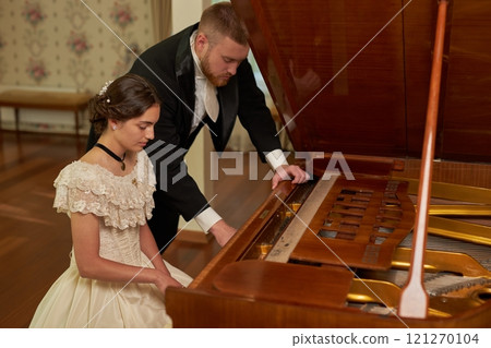 Side view portrait of classic young gentleman playing piano with lady in formal classical setting, copy space 121270104