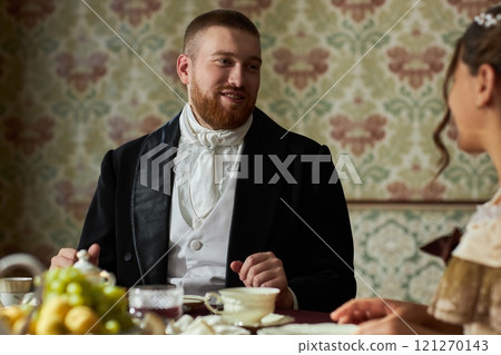 Portrait of bearded young gentleman smiling at young lady during elegant tea party in vintage setting Portrait of bearded young gentleman smiling at young lady during elegant tea party in vintage setting 121270143