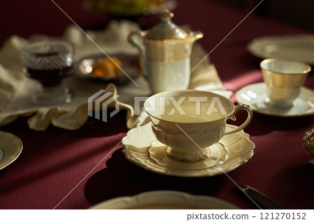 Close up of elegant porcelain tea cup with saucer on crimson tablecloth illuminated by natural light, copy space Close up of elegant porcelain tea cup with saucer on crimson tablecloth illuminated by natural light, copy space 121270152