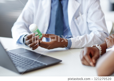 Cropped shot of African American doctor holding green pill bottle while explaining treatment to patient at desk in medical clinic, copy space 121270520