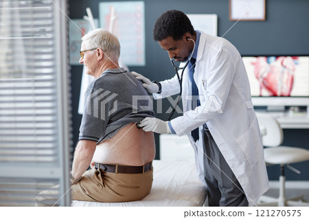 Side view of African American doctor using stethoscope and listening to heart and breathing while examining senior patient during checkup in clinic, copy space 121270575