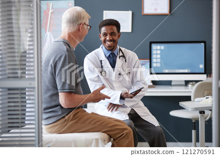Waist up portrait of smiling African American doctor wearing lab coat and holding tablet while talking to senior patient during consultation in clinic, copy space 121270591