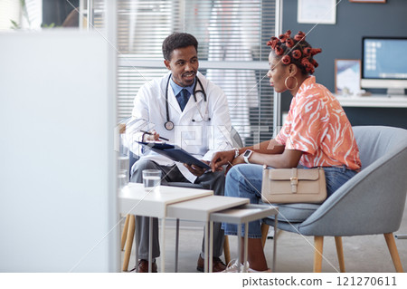 Full length portrait of African American doctor wearing lab coat and holding clipboard while discussing treatment with young woman during consultation in medical clinic setting Full length portrait of African American doctor wearing lab coat and holding clipboard while discussing treatment with young woman during consultation in medical clinic setting 121270611