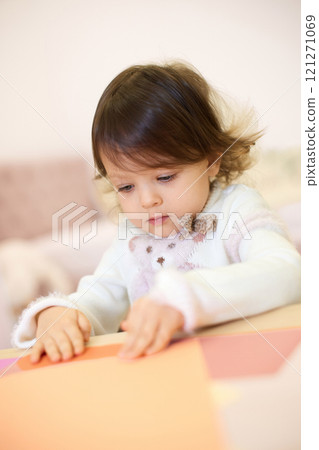 little child girl holding multi-color paper while sitting by table at home 121271069