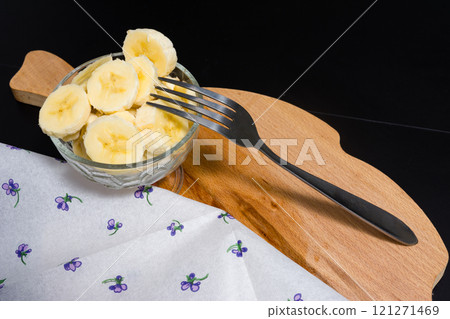Sliced banana in a glass bowl with a fork on a wooden board. 121271469