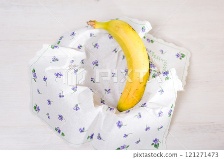 Ripe banana nestled in a floral patterned napkin on a white wooden table. 121271473