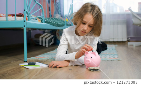 A young girl lies on her bedroom floor carefully placing coins into a piggy bank, learning about personal finance and the importance of saving 121273095