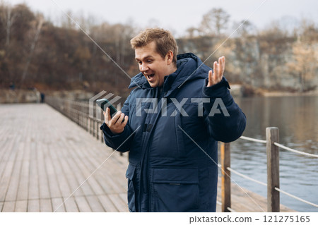Male standing on the lake wooden pier. Happy man using phone, walking near river. Phone Communication. Happy won the game, lottery. Human emotions, facial expression concept 121275165