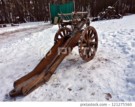 Antique cannon on a wooden gun carriage with wheels. Winter in the snow in the park against the background of a cart and trees Antique cannon on a wooden gun carriage with wheels. Winter in the snow in the park against the background of a cart and trees 121275560
