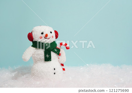 snowman with red earmuffs, a green scarf, and a candy cane stands on snowy ground against a light blue background snowman with red earmuffs, a green scarf, and a candy cane stands on snowy ground against a light blue background 121275690