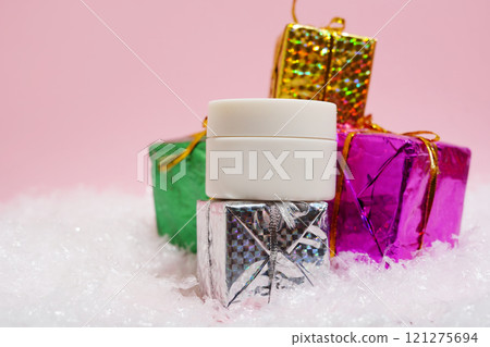 A white cosmetic jar is displayed on shiny, colorful gift boxes surrounded by artificial snow against a soft pink background A white cosmetic jar is displayed on shiny, colorful gift boxes surrounded by artificial snow against a soft pink background 121275694