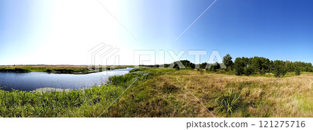 Panorama of the river and meadows with herbs against the blue sky. Still green grass and river vegetation and withered dry grasses in the pasture. 121275716