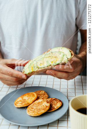 man has breakfast at home in kitchen. plate with avocado toast, pancakes and black coffee man has breakfast at home in kitchen. plate with avocado toast, pancakes and black coffee 121275772