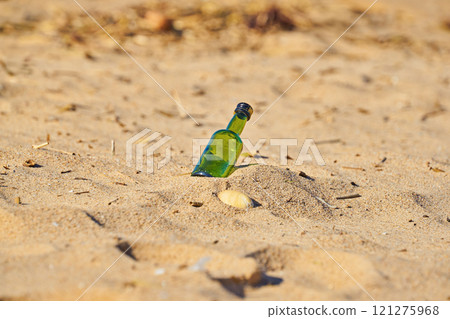 Green glass bottle tilted and partially buried in sand on sunny beach, surrounded by scattered natural debris and shells 121275968