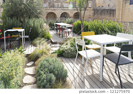 Modern outdoor dining area featuring colorful chairs and white tables surrounded by lush greenery on wooden deck with stone building backdrop 121275970