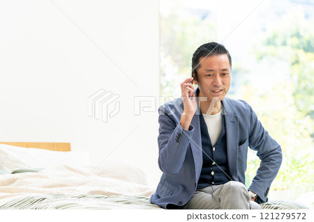 Middle-aged man sitting on bed and talking on smartphone Middle-aged man sitting on bed and talking on smartphone 121279572