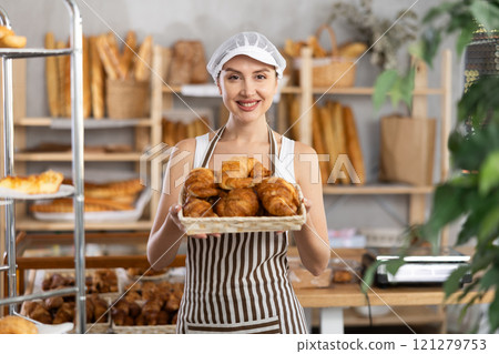Young saleswoman displays croissants in square wicker basket 121279753