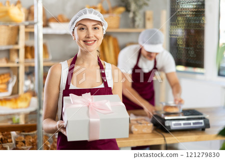 Cheerful female baker holding gift box with baked treats Cheerful female baker holding gift box with baked treats 121279830