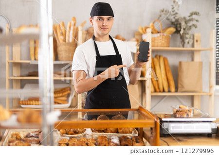 Guy seller in bakery hold out hand with phone, points at screen Guy seller in bakery hold out hand with phone, points at screen 121279996