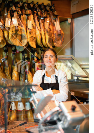 Portrait of positive female butcher on background of rack with hanging various jamon 121280023