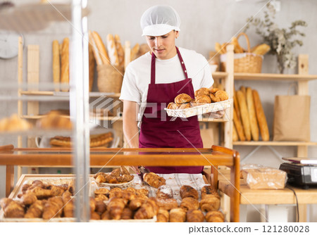 Young man employee puts croissants in window, arranges display of goods at bakery. 121280028
