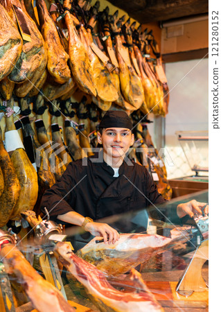 Male seller cutting slices from whole leg of jamon in meat store Male seller cutting slices from whole leg of jamon in meat store 121280152