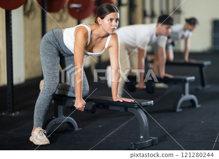 Athletic young woman pumping her biceps with a dumbbell while leaning on bench Athletic young woman pumping her biceps with a dumbbell while leaning on bench 121280244