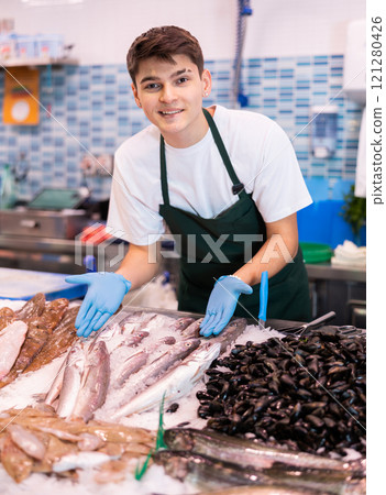 Seller in apron standing near counter offering fresh fish striped red mullet Seller in apron standing near counter offering fresh fish striped red mullet 121280426