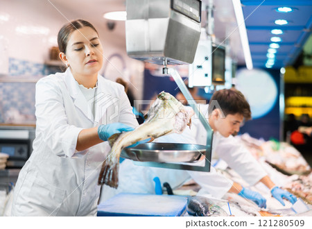 Female seller in fish department of supermarket weighs atlantic cod fish on scale 121280509