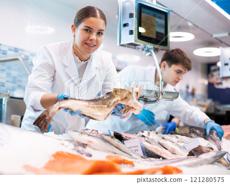 Positive saleswoman holding cod fish in fish store 121280575