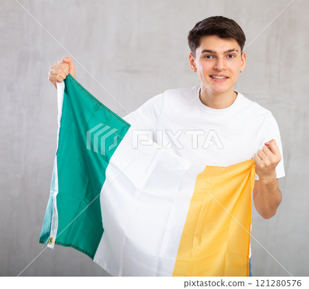 Enthusiastic young man holding flag of Ireland against unicoloured background Enthusiastic young man holding flag of Ireland against unicoloured background 121280576