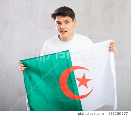 Reliant positive young man holds big national flag of Algeria with confident smile showing teeth. 121280577