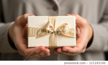 Hands holding small gift box with gold ribbon bow, close-up, beige background, special occasion, celebration, surprise, festive, elegant, wrapped package, thoughtful gesture, love 121283140