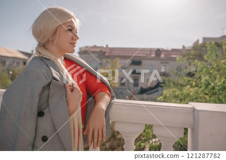 A blonde woman wearing a red shirt and gray jacket stands on a balcony. She is wearing ripped jeans and has her hands in her pockets. A blonde woman wearing a red shirt and gray jacket stands on a balcony. She is wearing ripped jeans and has her hands in her pockets. 121287782