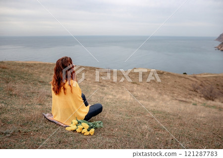 A woman sits on the grass with a bouquet of yellow flowers in her lap. She is looking out at the ocean, taking in the view. Concept of peace and tranquility. 121287783