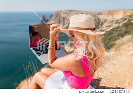 A woman is sitting on a rocky beach with a laptop in front of her. She is wearing a pink top and a straw hat. Concept of relaxation and leisure. A woman is sitting on a rocky beach with a laptop in front of her. She is wearing a pink top and a straw hat. Concept of relaxation and leisure. 121287803
