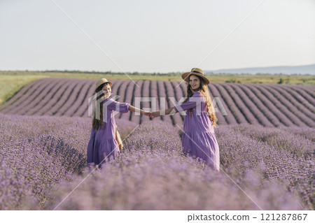 Mom and daughter are running through a lavender field dressed in purple dresses, long hair flowing and wearing hats. The field is full of purple flowers and the sky is clear. Mom and daughter are running through a lavender field dressed in purple dresses, long hair flowing and wearing hats. The field is full of purple flowers and the sky is clear. 121287867