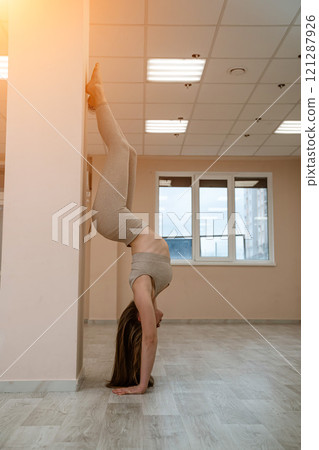 The girl is training in the yoga studio. Dressed in a beige tracksuit doing a handstand against the wall. Healthy lifestyle and yoga concept. The girl is training in the yoga studio. Dressed in a beige tracksuit doing a handstand against the wall. Healthy lifestyle and yoga concept. 121287926