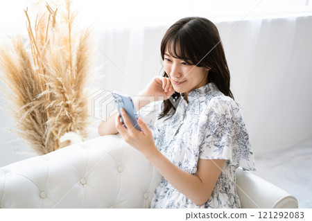 A young woman with long black hair sitting on a white sofa and holding a smartphone 121292083