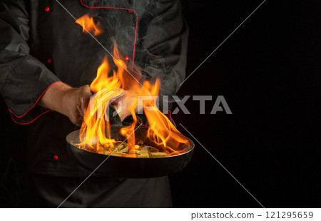 In a modern kitchen, a skilled chef expertly tosses sizzling ingredients in a skillet, igniting a spectacular display of flames. The dramatic fire contrasts beautifully with the dark background 121295659