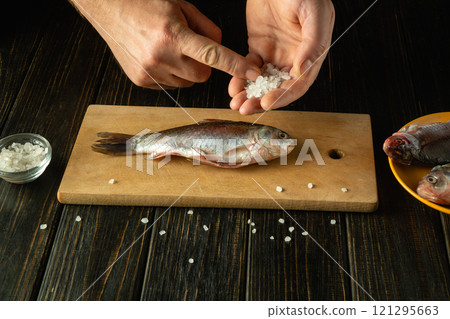Skilled hands sprinkle coarse salt over a fresh fish on a wooden cutting board, setting the stage for a flavorful dish. Nearby, additional fish await their turn in the kitchen Skilled hands sprinkle coarse salt over a fresh fish on a wooden cutting board, setting the stage for a flavorful dish. Nearby, additional fish await their turn in the kitchen 121295663