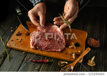 Hands skillfully season a fresh cut of meat with aromatic herbs and spices on a wooden cutting board. The warm ambiance of the rustic kitchen adds to the culinary creativity 121295683