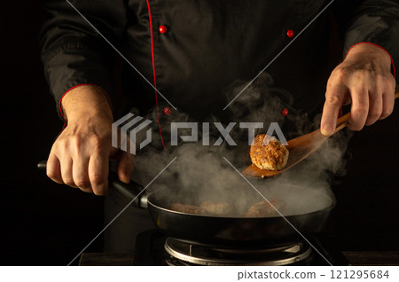 A chef expertly flips golden-brown patties in a pan filled with steam, showcasing their cooking technique. The dark background amplifies the intensity of this culinary moment, highlighting artistry 121295684