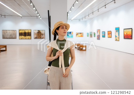 An above-the-knee portrait of a young elegantly dressed woman in a straw hat admiring the exhibition of paintings in the gallery. Contemporary art at the Museum. Cultural education concept 121298542