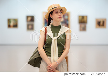 Mid shot of a young Caucasian woman in a straw hat and elegant clothes contemplating modern art in the gallery. Copy space. The concept of the Museum Day Mid shot of a young Caucasian woman in a straw hat and elegant clothes contemplating modern art in the gallery. Copy space. The concept of the Museum Day 121298544