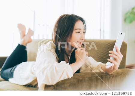 A woman lying on the sofa and looking at her smartphone A woman lying on the sofa and looking at her smartphone 121299738