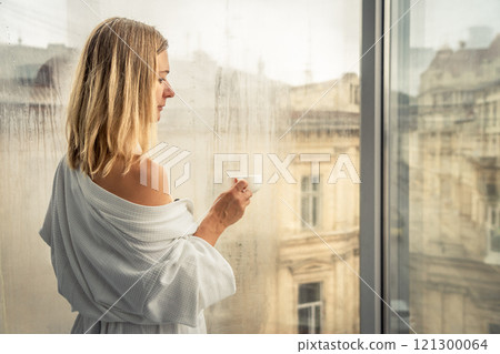 Woman in elegant robe drinking coffee in hotel room and standing near window 121300064