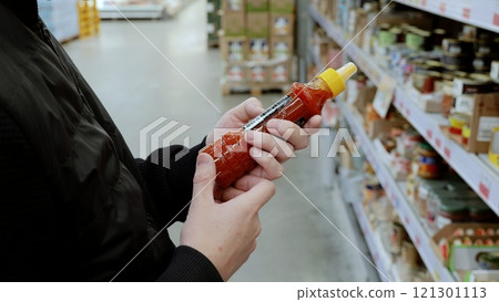 Shopper in a crowded supermarket aisle carefully examines a hot sauce bottle, trying to decide if it's worth purchasing among the variety of spices on the shelves Shopper in a crowded supermarket aisle carefully examines a hot sauce bottle, trying to decide if it's worth purchasing among the variety of spices on the shelves 121301113